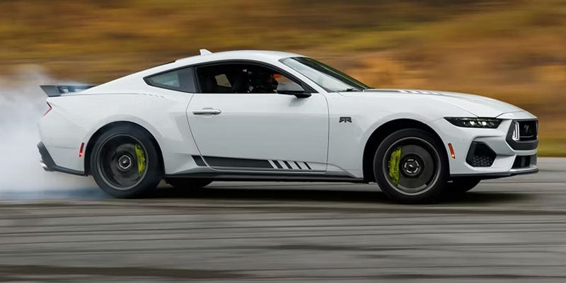 White Ford Mustang drifting or doing a burnout, with smoke from the rear tire and motion blur.