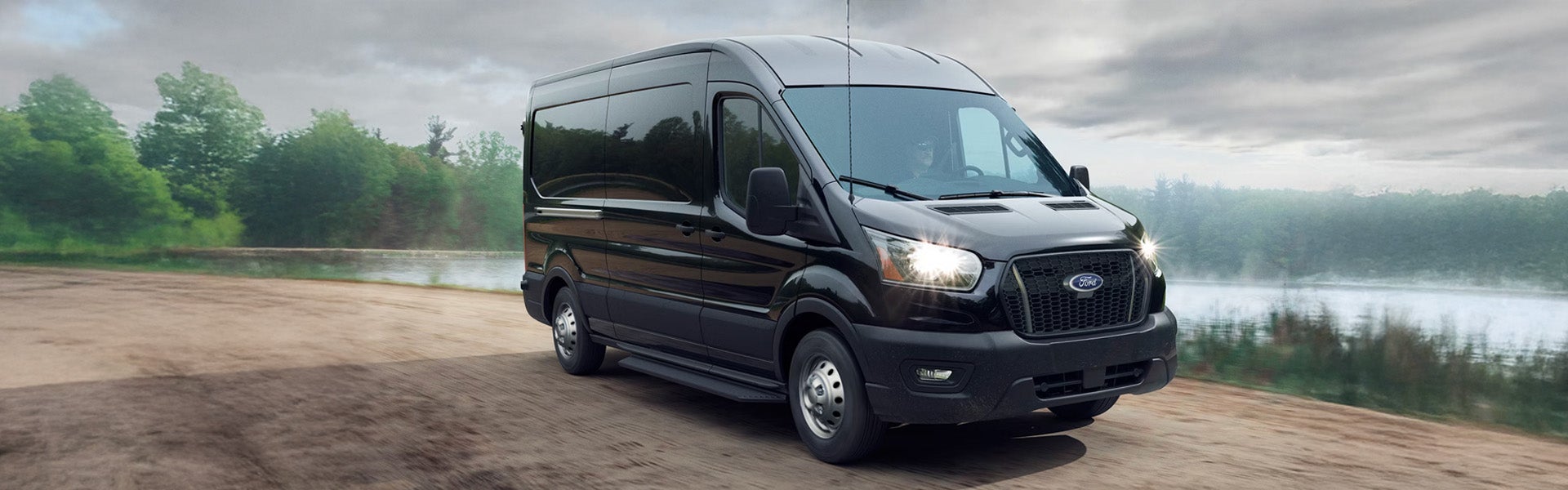 Black Ford Transit van on a dirt path with headlights on, next to a lake, under a cloudy sky.